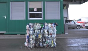 Bale of compressed mixed plastic waste secured with baler wire positioned in front of a recycling facility control building.
