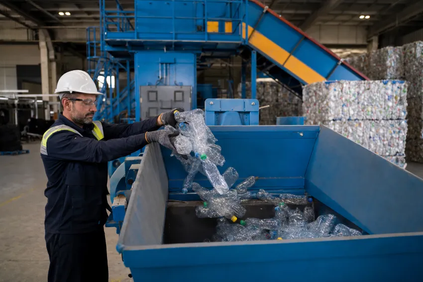 Warehouse operator loading clear PET plastic bottles into the hopper of a vertical plastic baler machine.