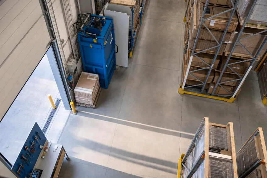 Overhead view of a compact vertical cardboard baler machine sitting flush against a wall in a clean retail warehouse aisle.